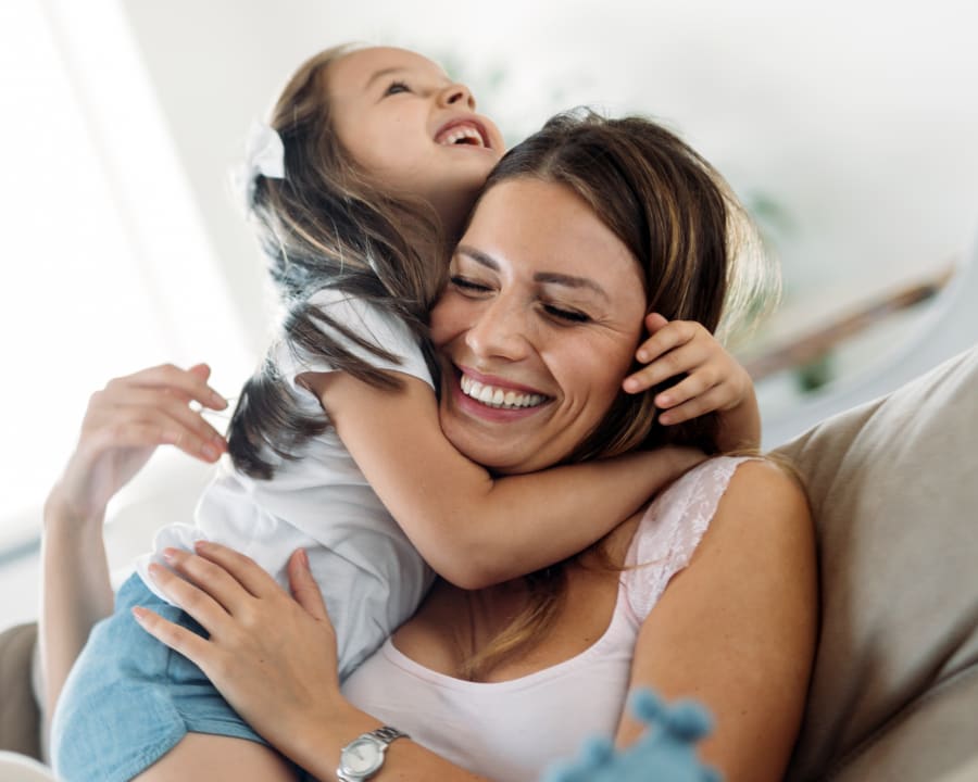 A mother on a couch smiles as her young daughter climbs on top of her.
