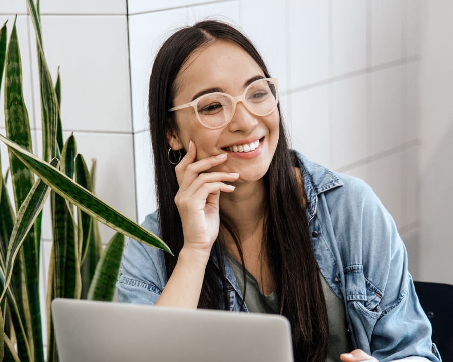 A woman with glasses sits at a table in a coffee shop in front of a laptop smiling.