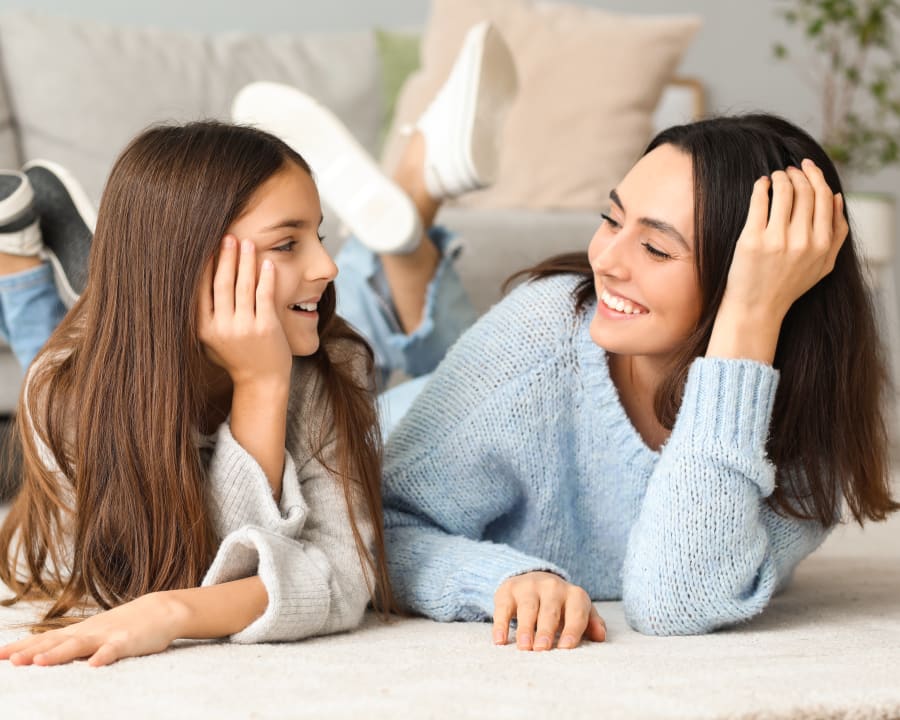 A woman and girl lay on the stomachs on the floor in front of a sofa smiling at eachother.