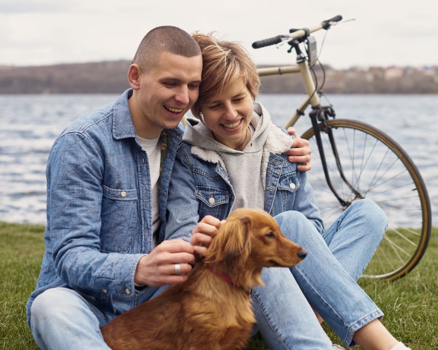 A man and woman sit on the ground outside next a river. They are in front of a bicycle and petting a small brown dog.