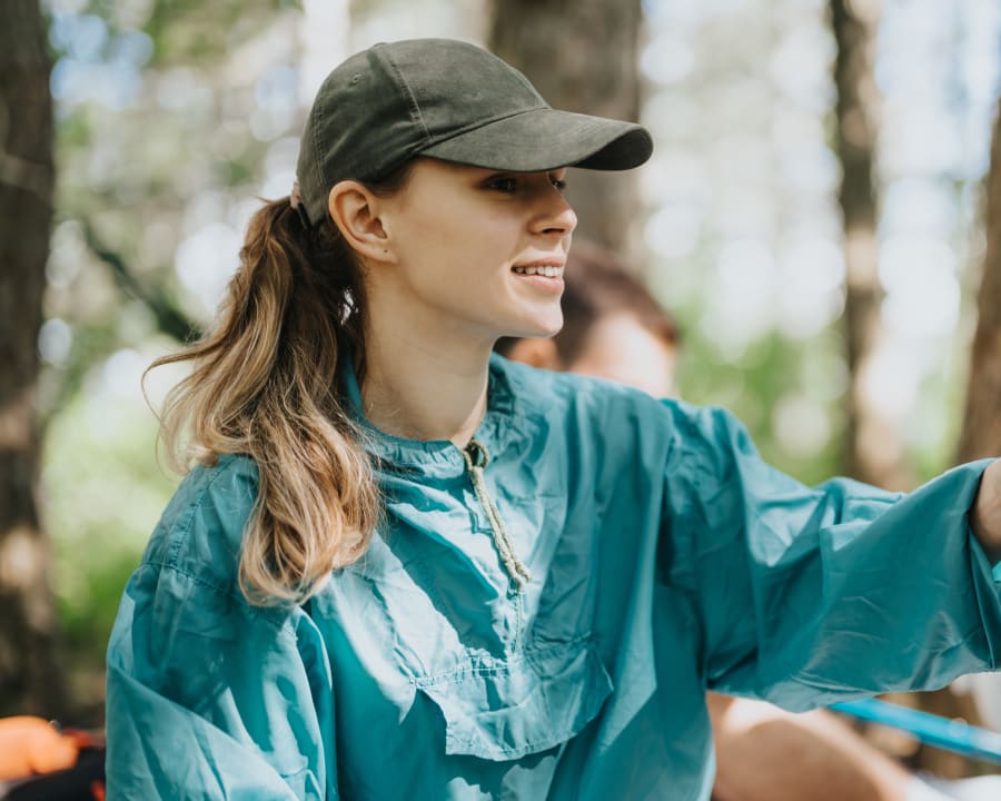 A young woman wearing a blue windbreaker with a pony tail in a black baseball cap is sitting outside with her friends.