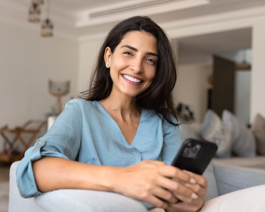 A woman with long hair sits on the couch and smiles while holding her phone with both hands.