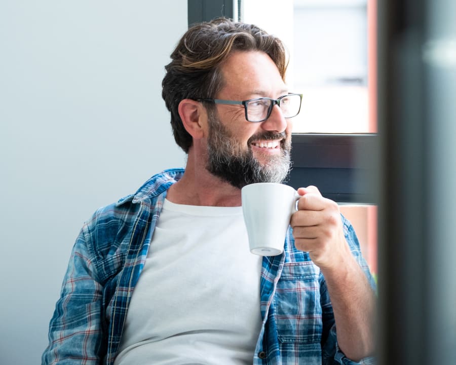 A man with short hair and glasses wears a blue plaid shirt and is holding a mug up while smiling and looking off to the side.